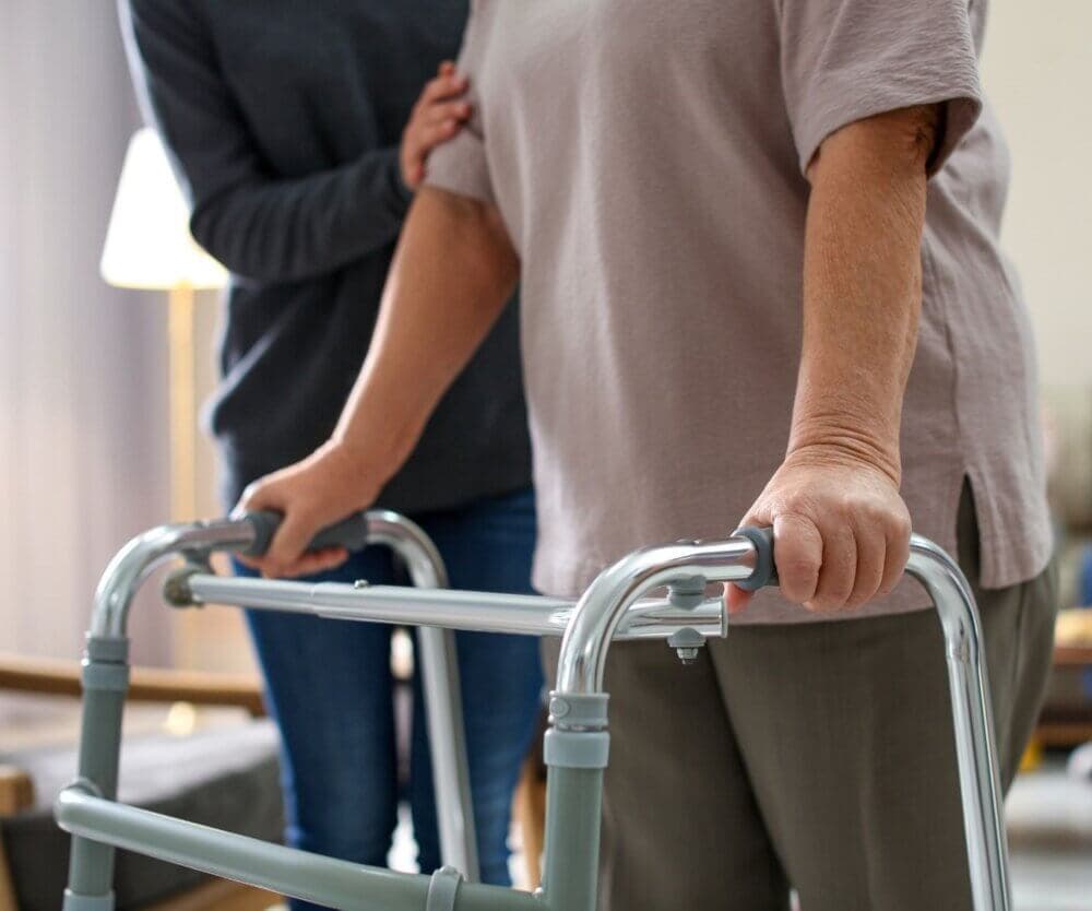 A person in a pink shirt using a walker, with another person in a gray shirt offering support by holding their arm. - Home Instead