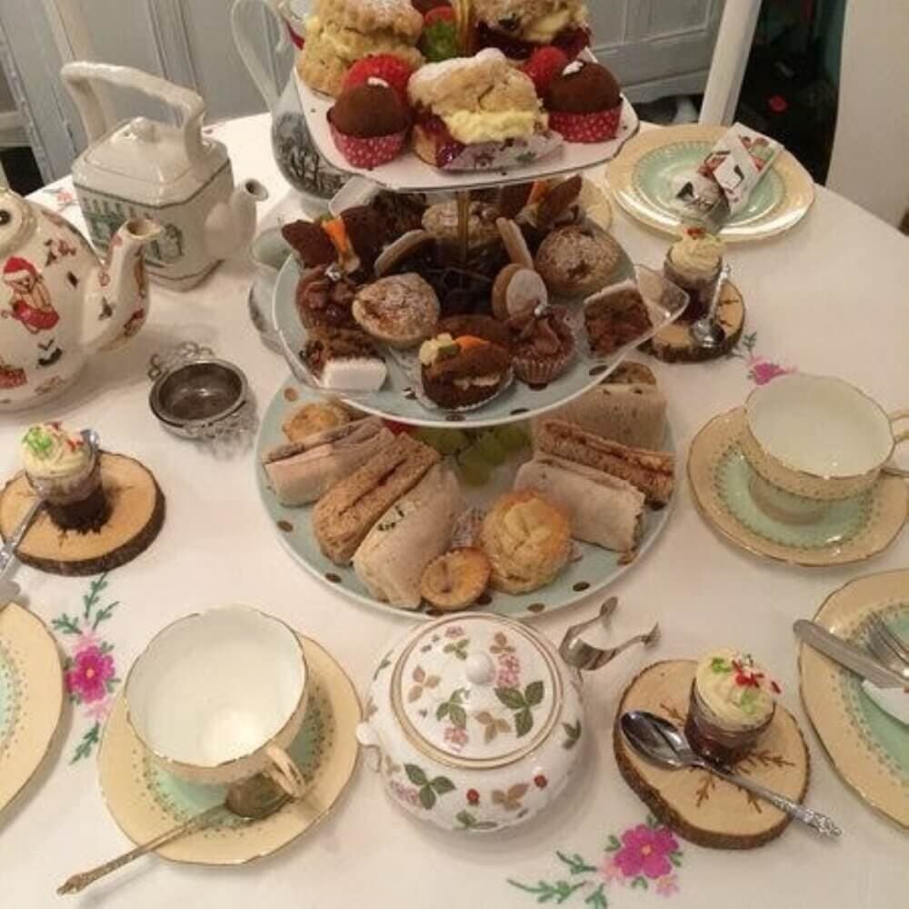 A table set for tea with assorted pastries, sandwiches, and tea cups on a floral tablecloth. - Home Instead