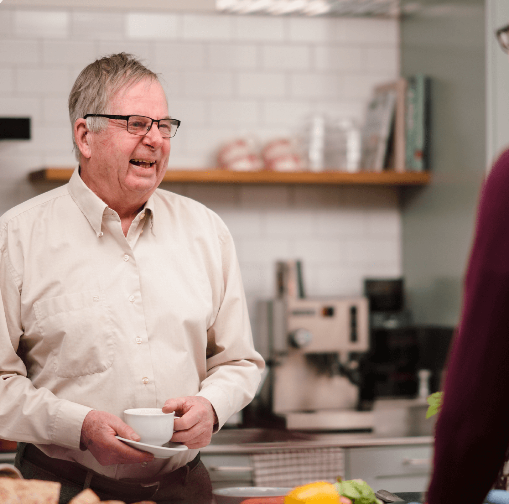 An elderly man holding a cup and smiling while talking to someone in a kitchen setting. - Home Instead