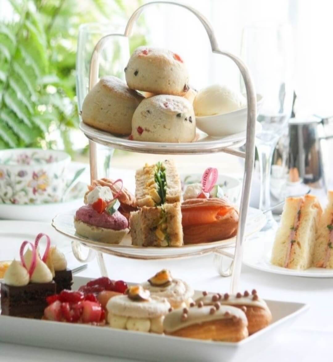 A tiered tray with scones, assorted sandwiches, and pastries. There are also plates and teacups on a table set for tea. - Home Instead