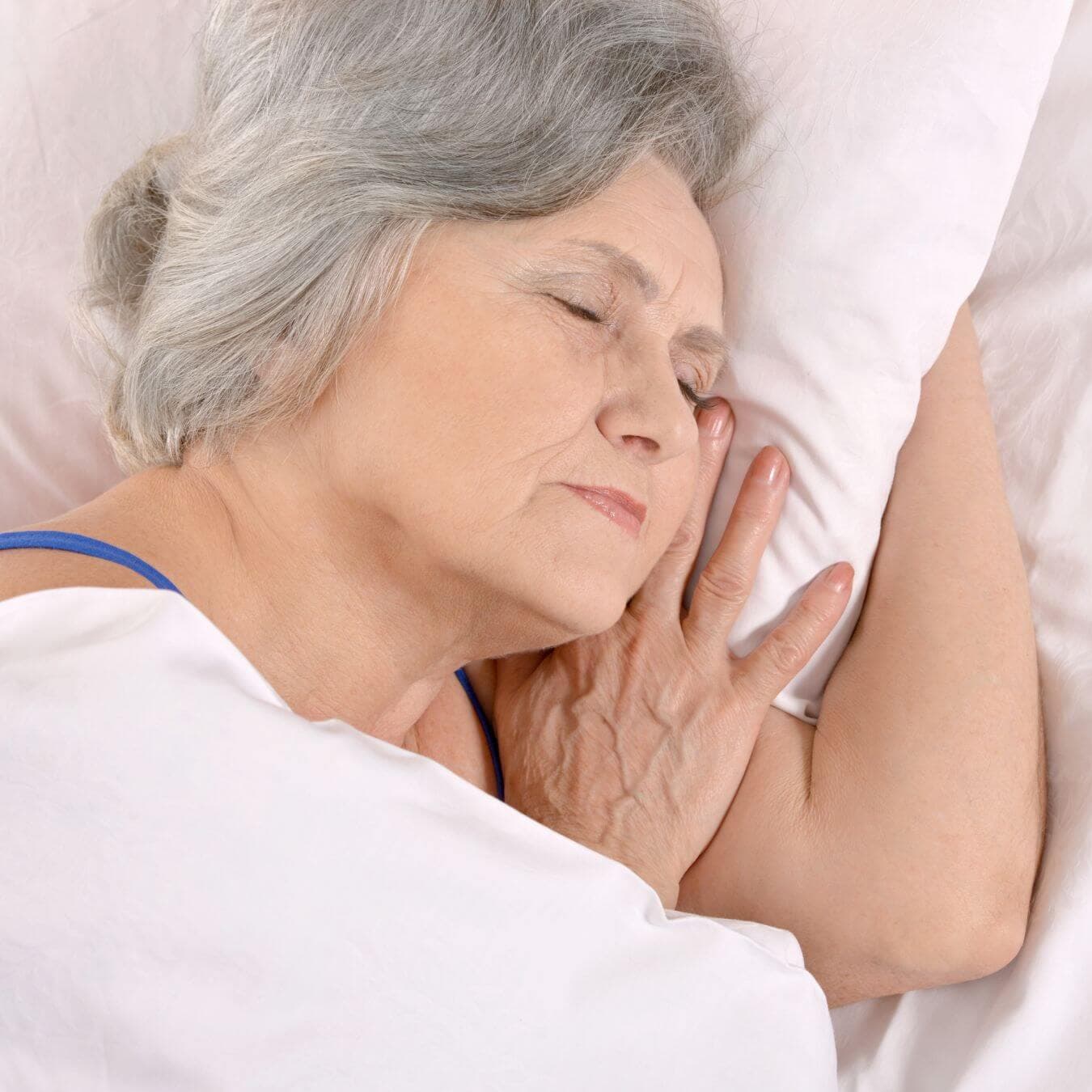 Elderly woman with gray hair sleeping peacefully on her side in bed, covered with a white sheet. - Home Instead