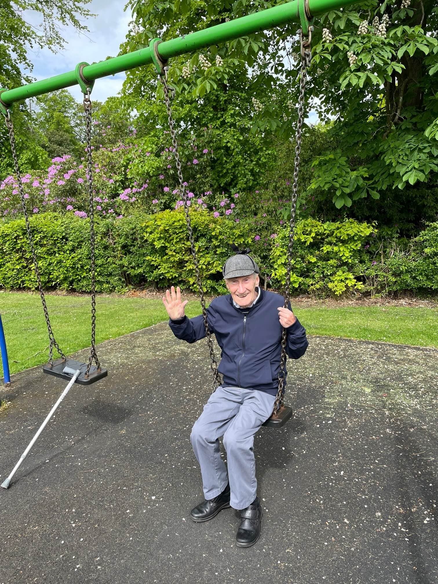 Elderly man in a jacket and cap, sitting on a swing in a park, smiling and waving, with greenery in the background. - Home Instead