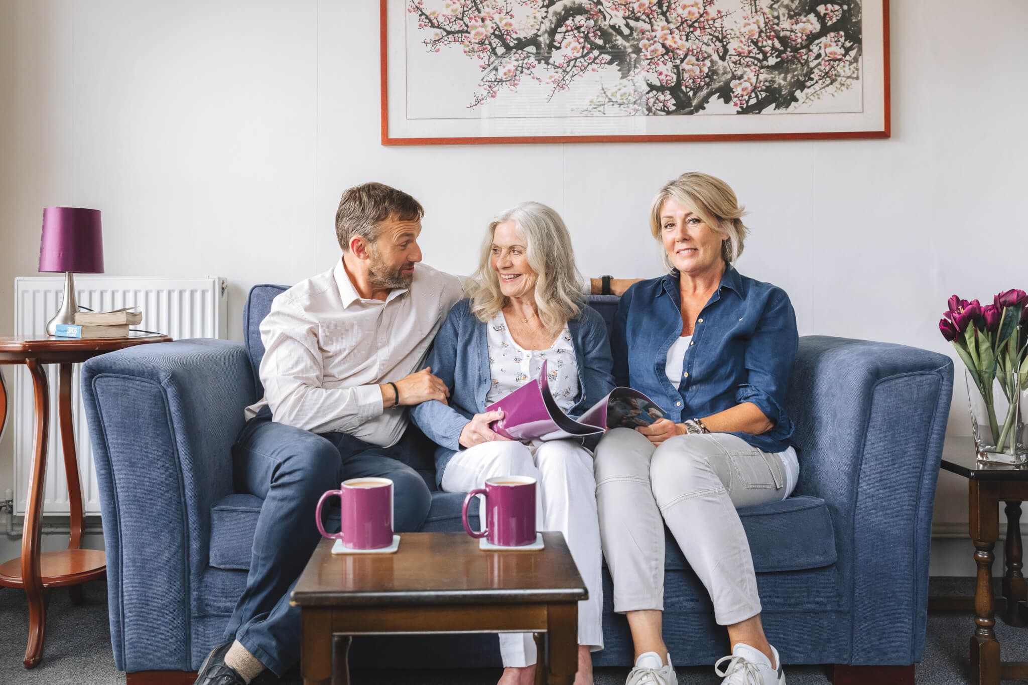 Three people sitting on a blue couch, smiling, with two mugs on the table and a floral painting in the background. - Home Instead