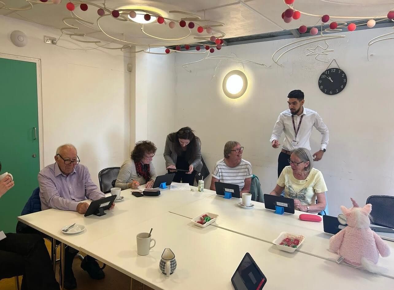 A group of elderly people and two facilitators engage with tablets during a tech workshop in a decorated room. - Home Instead