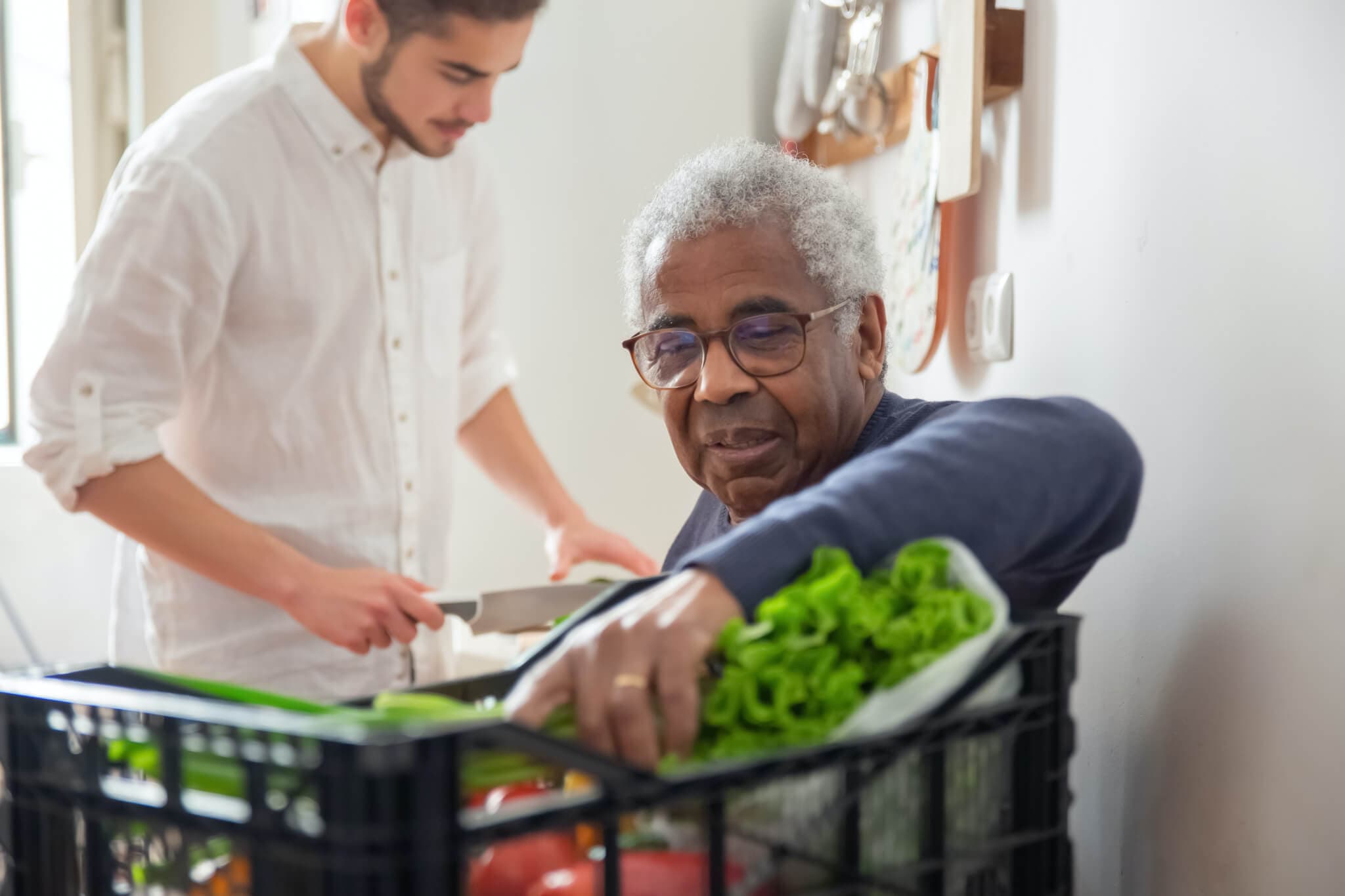Elderly man picks vegetables from a crate while a younger man chops vegetables in the background. - Home Instead