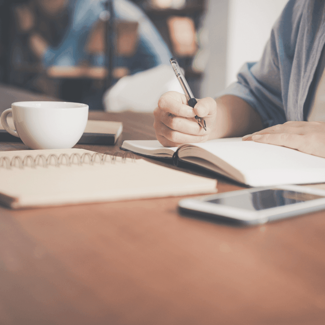 Person writing in a notebook on a wooden table, with a smartphone, coffee cup, and another book nearby. - Home Instead
