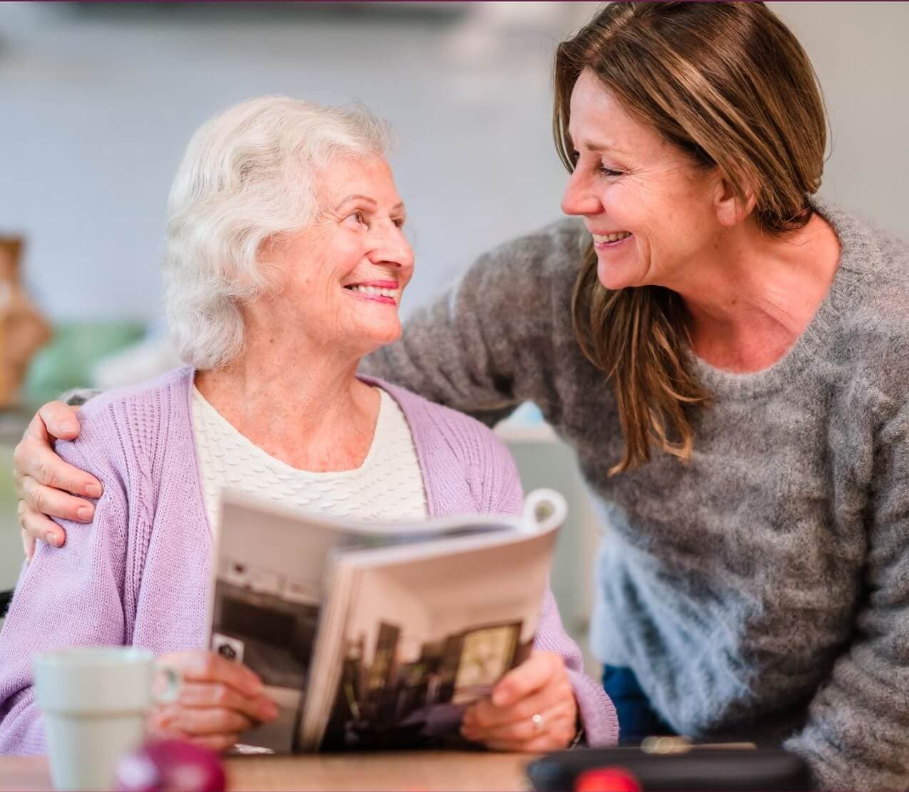 An elderly woman and a younger woman smile at each other; one holds a magazine, the other has her arm around her. - Home Instead