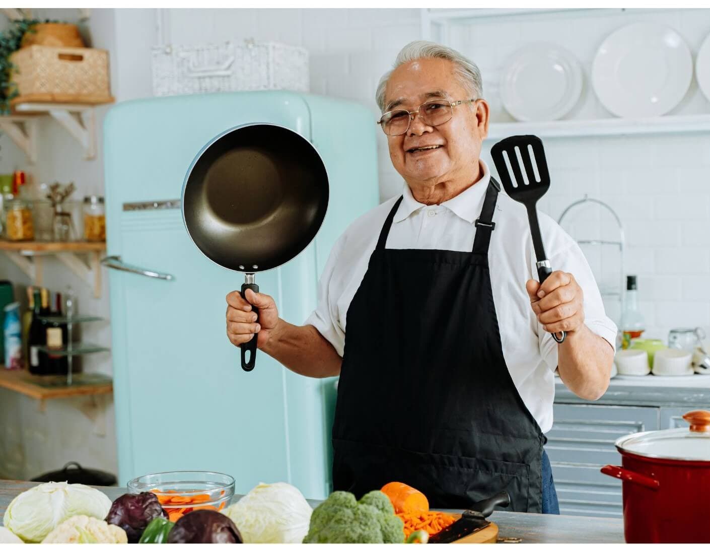 Smiling senior man in an apron holding a frying pan and spatula in a kitchen with vegetables on the counter. - Home Instead