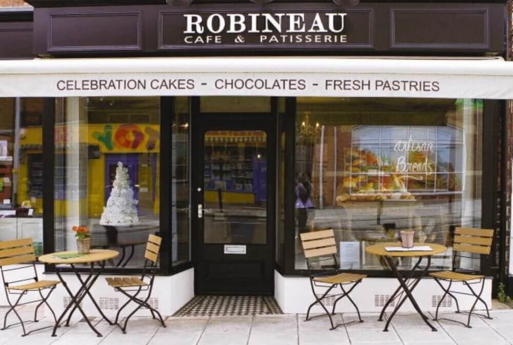 Exterior of Robineau Café & Patisserie with outdoor seating and a display of celebration cakes and pastries in the window. - Home Instead