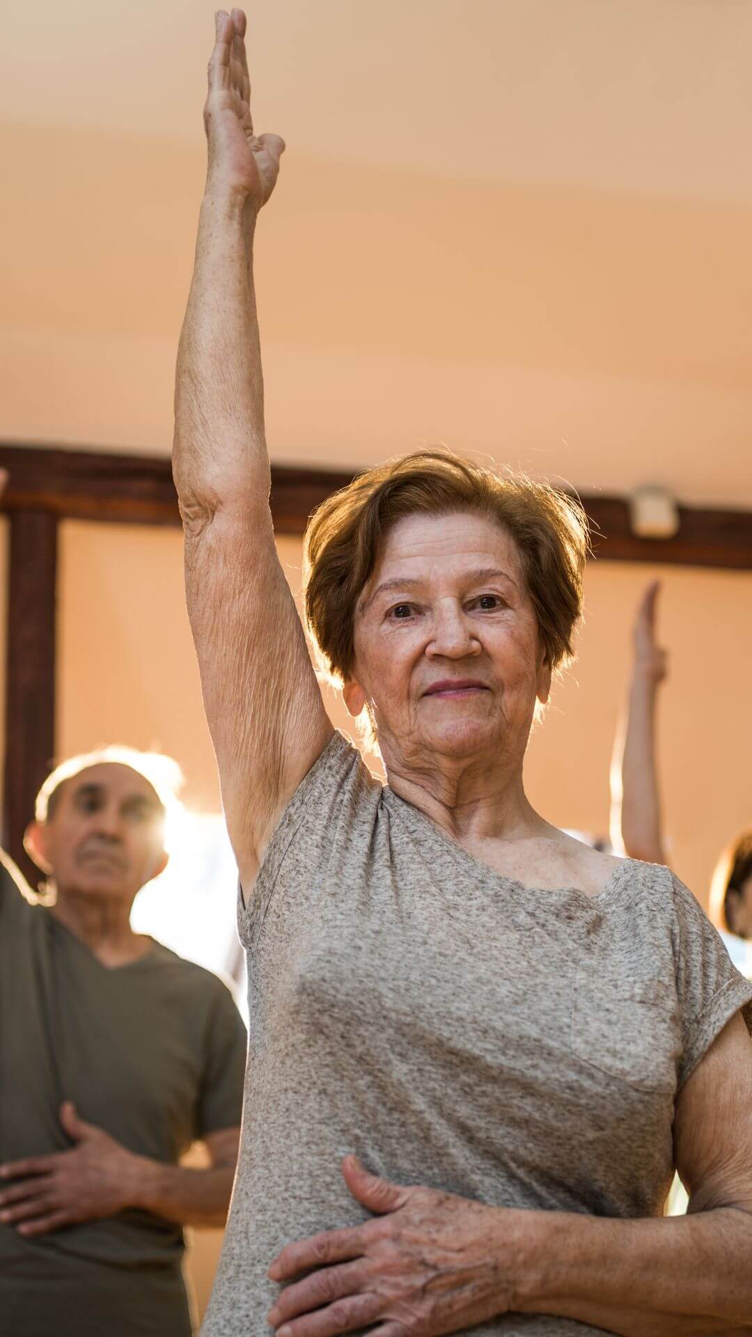 Elderly woman in a gray shirt raises her arm during a group exercise class, with others in the background. - Home Instead