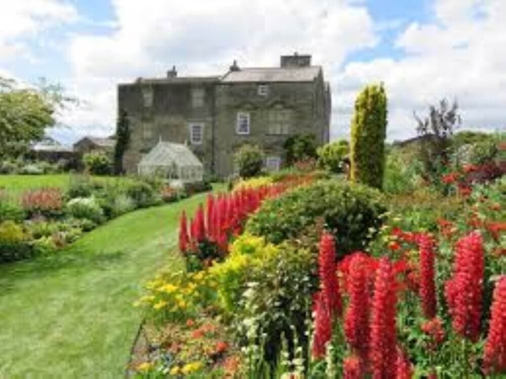 A stone house with a greenhouse and lush garden filled with vibrant flowers, including red lupines, under a partly cloudy sky. - Home Instead