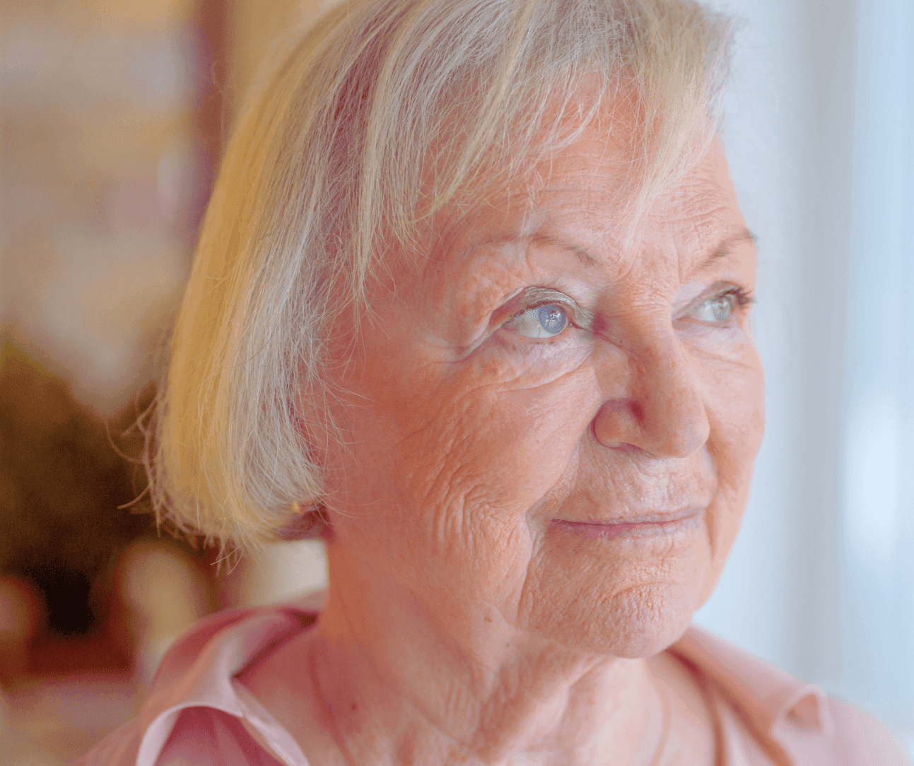 Elderly woman with short blonde hair and a thoughtful expression gazes to the side in front of a blurred background. - Home Instead