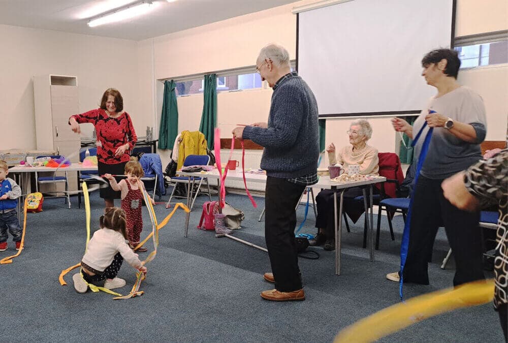 A group of people, young and old, play with ribbons in a community room with a screen and chairs in the background. - Home Instead
