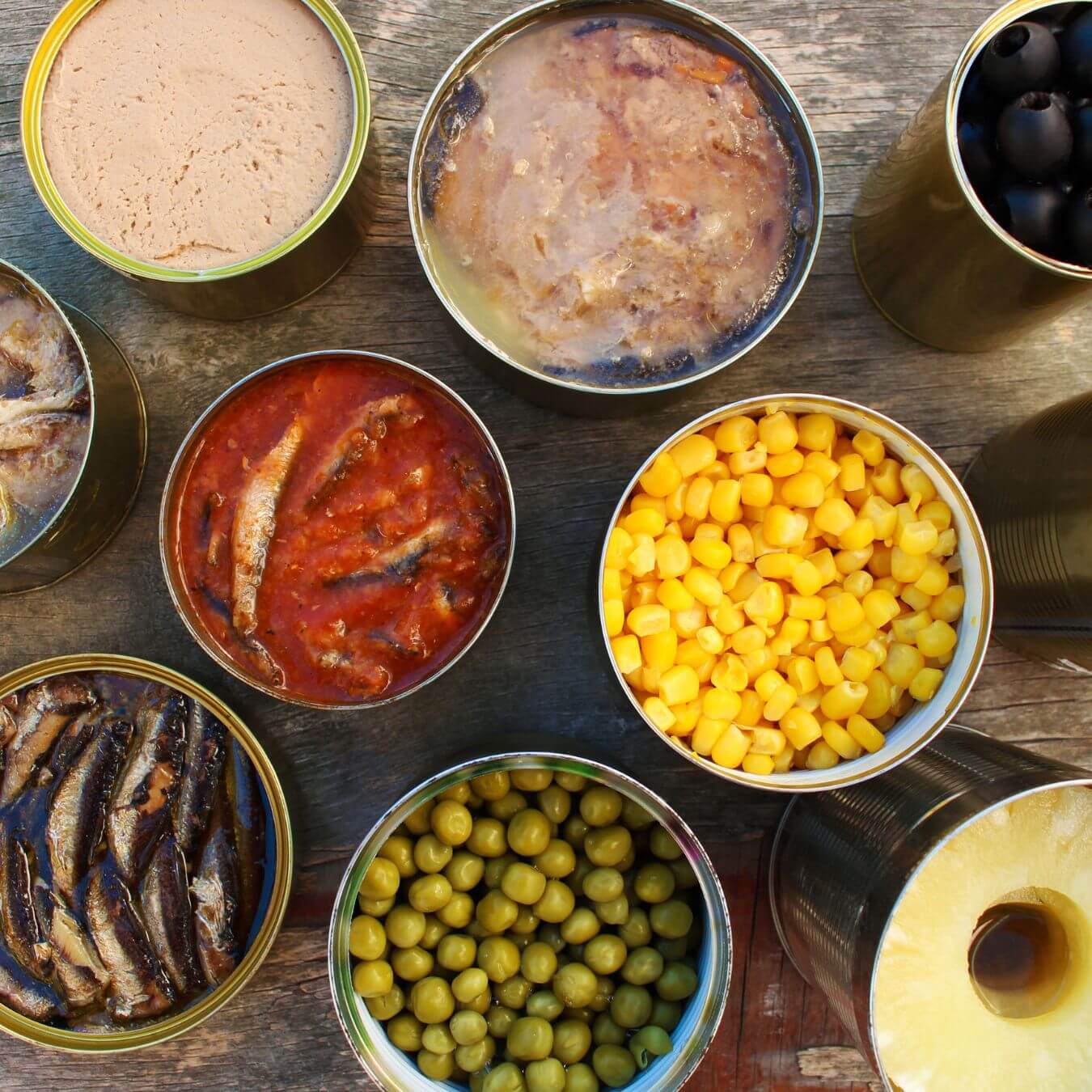 Various opened canned foods including fish, corn, peas, olives, pineapple, and meat on a wooden surface. - Home Instead