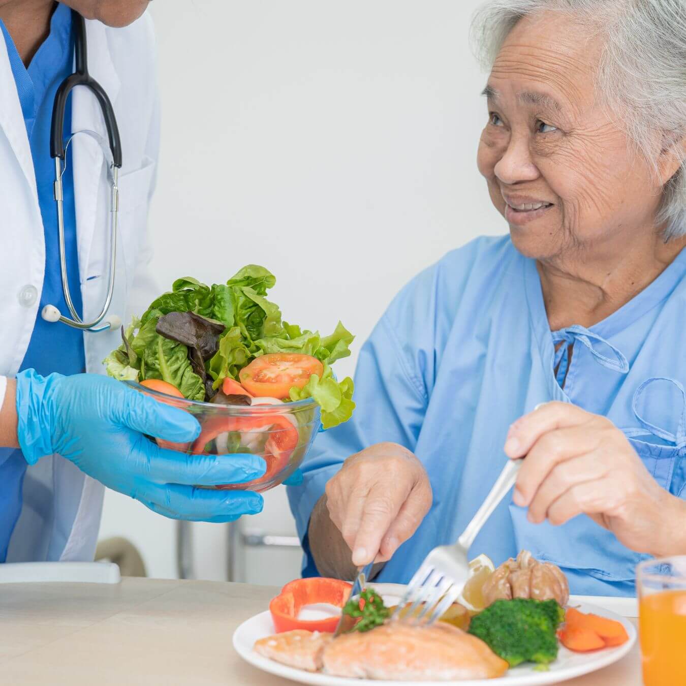 A doctor hands a salad to a smiling elderly woman eating a meal in a healthcare setting. - Home Instead