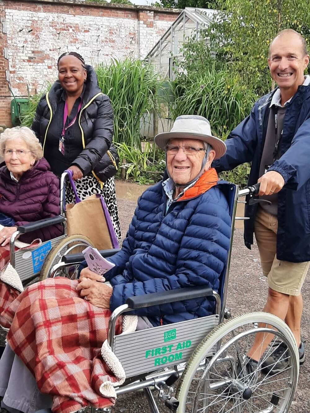 Two elderly people in wheelchairs, outside with two caregivers smiling, on a cloudy day. - Home Instead
