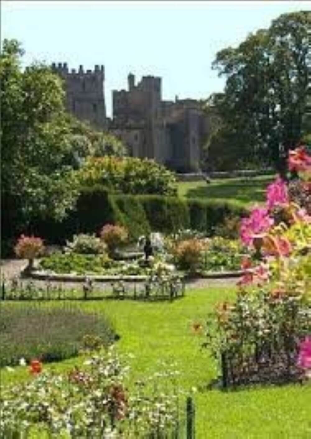 A lush garden with blooming flowers and hedges, featuring a stone castle in the background under a clear sky. - Home Instead