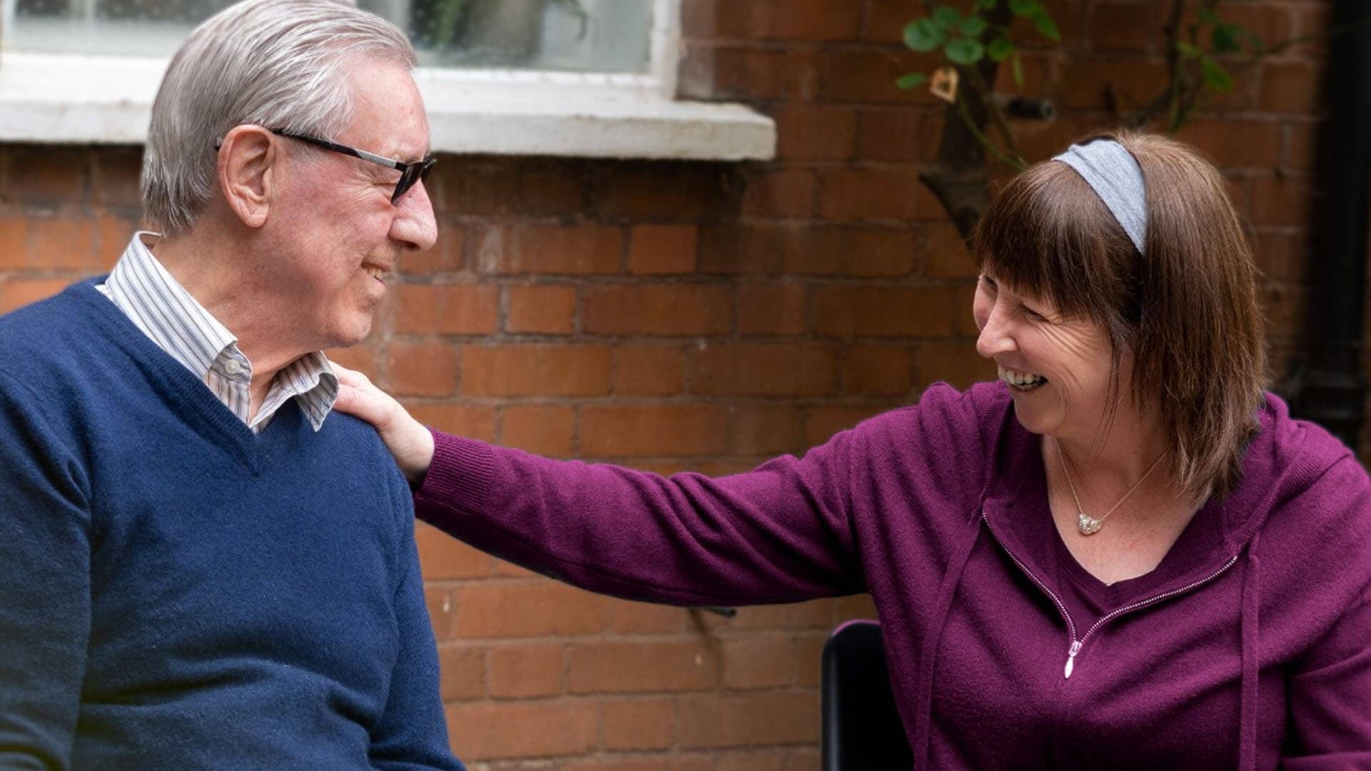 An older man and woman share a warm laugh outdoors, with the woman lightly touching the man's shoulder. - Home Instead