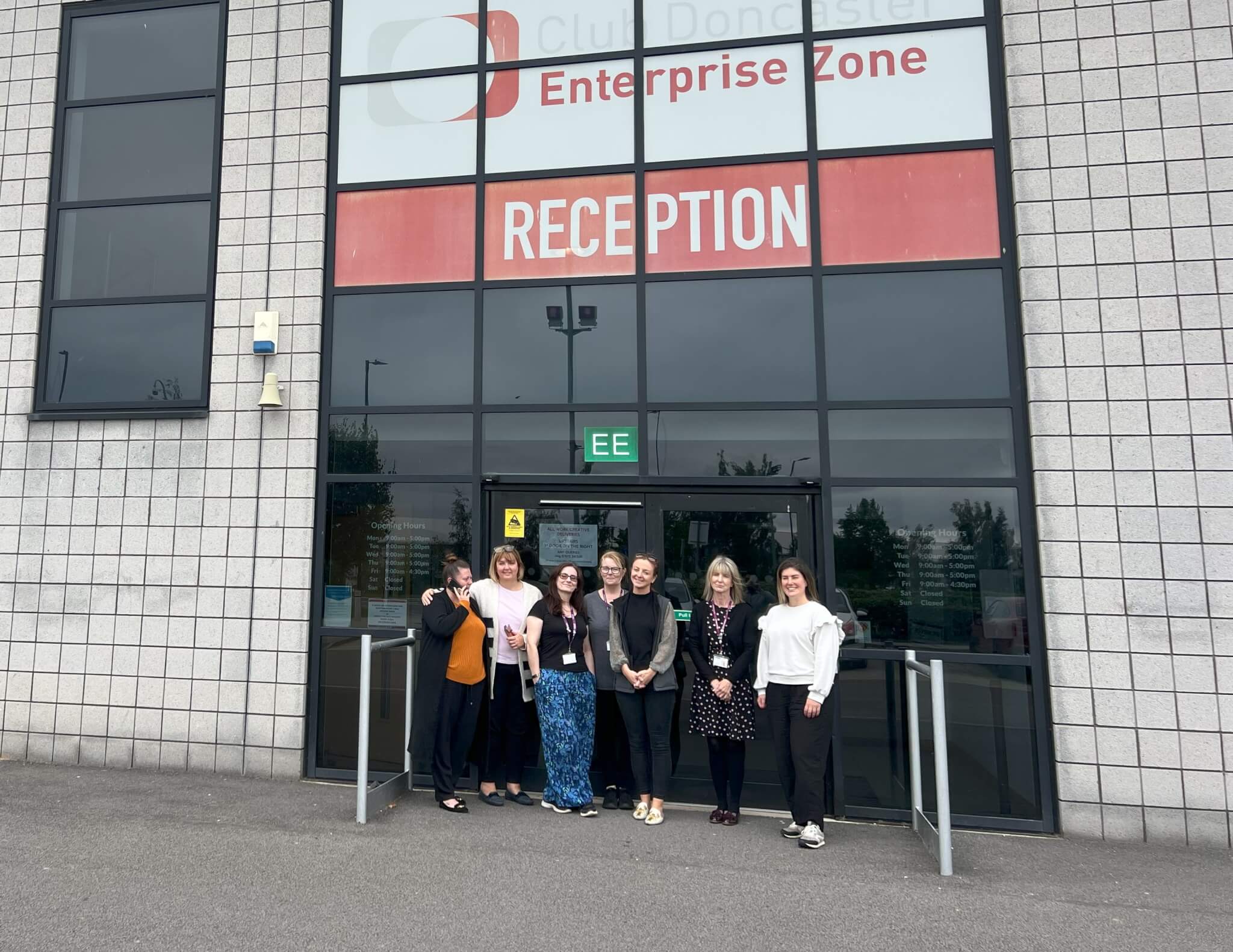 Seven people standing and posing for a photo in front of a building with a sign reading "RECEPTION" above the entrance. - Home Instead