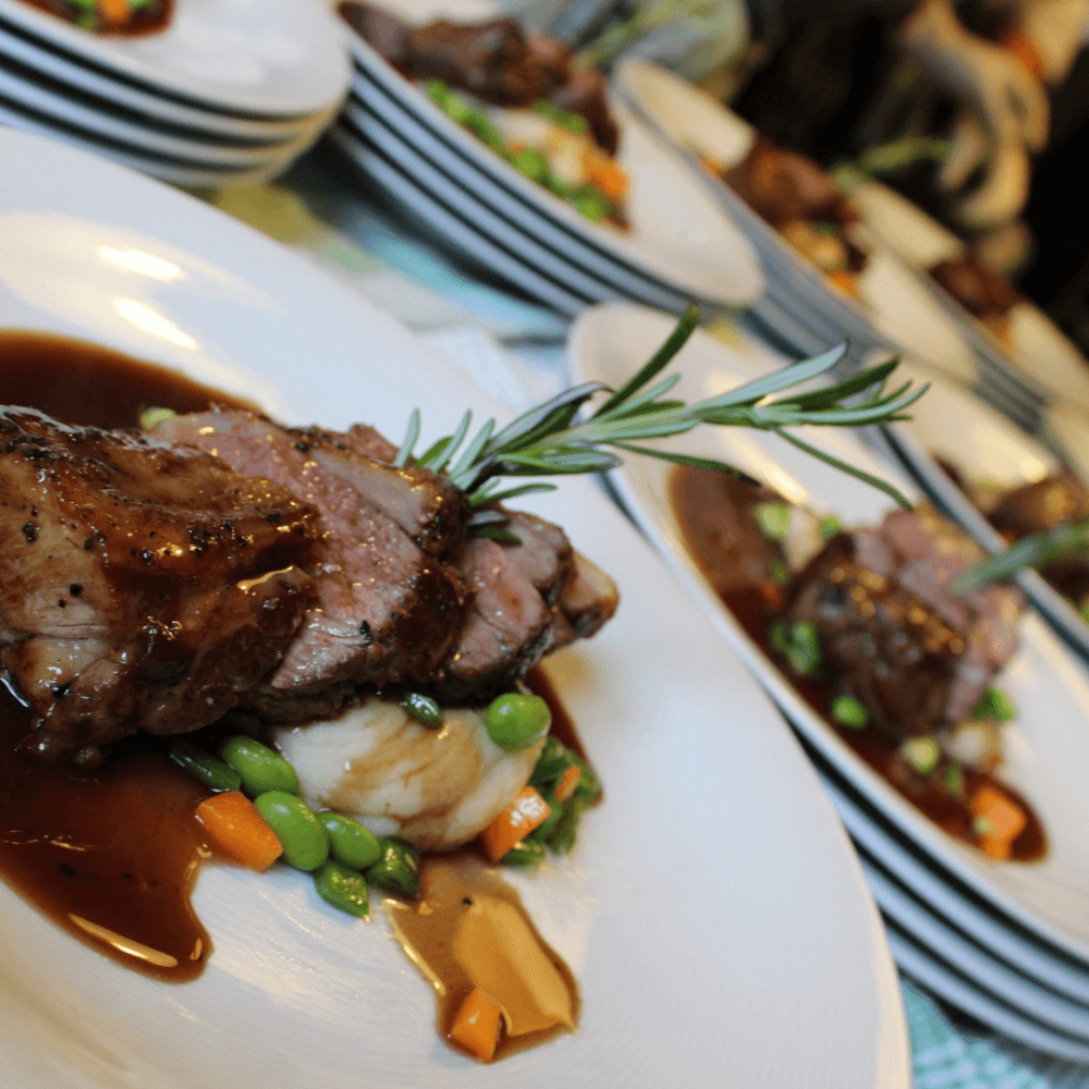 Sliced steak served on mashed potatoes, garnished with peas, carrots, and rosemary, with a brown sauce. - Home Instead