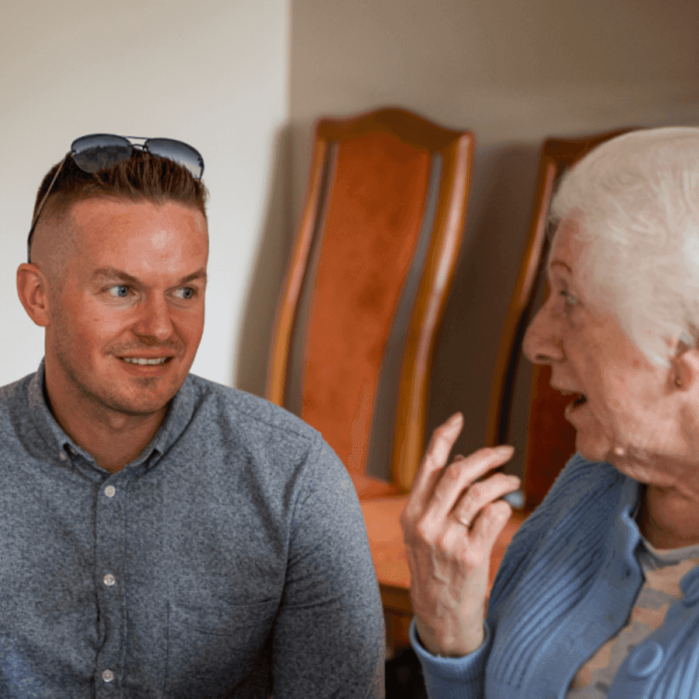 Man with sunglasses on his head conversing with an elderly woman in a blue sweater, wooden chairs in the background. - Home Instead