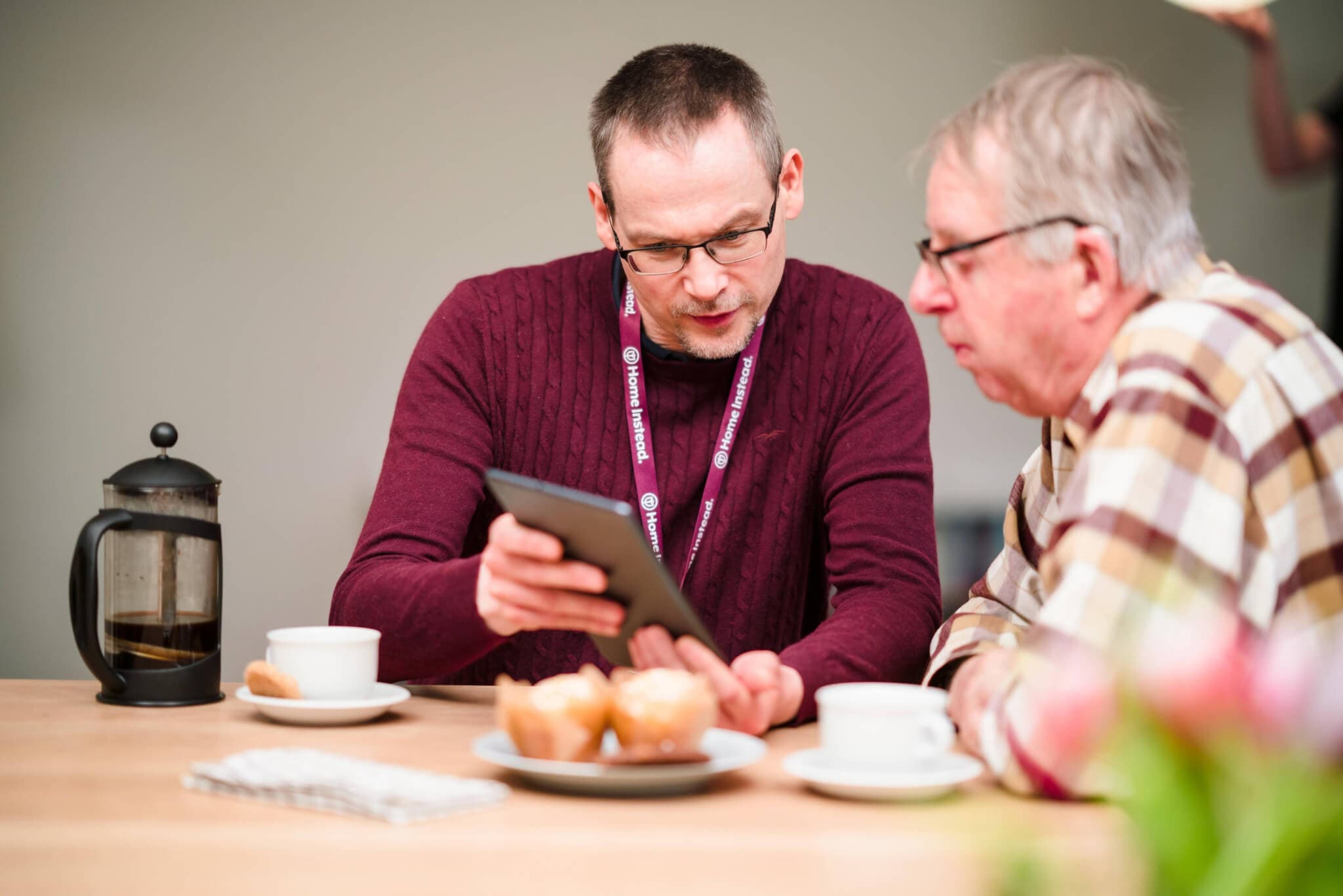 Two men sitting at a table with coffee and pastries, one showing the other something on a tablet. - Home Instead