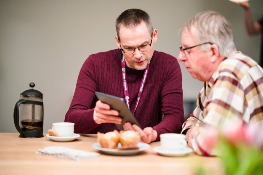 Two men at a table, one in a maroon sweater showing a tablet to the other in a plaid shirt, with coffee and pastries. - Home Instead