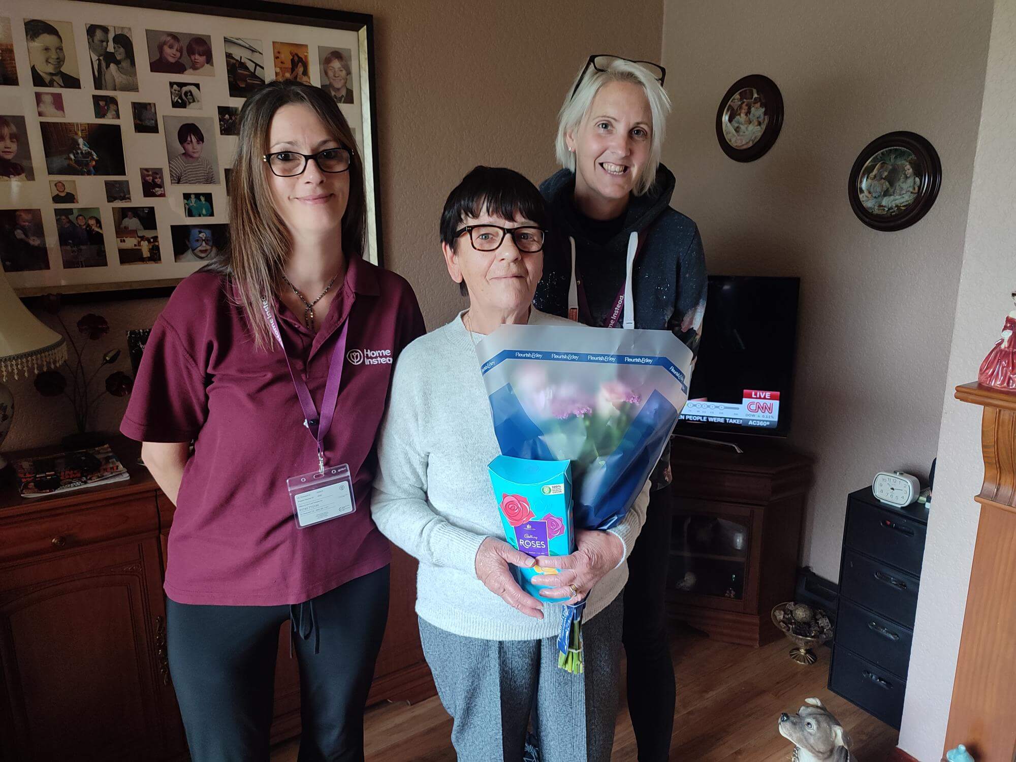 Three women standing together; middle one holding flowers inside a cozy room with picture frames and a dog statue. - Home Instead