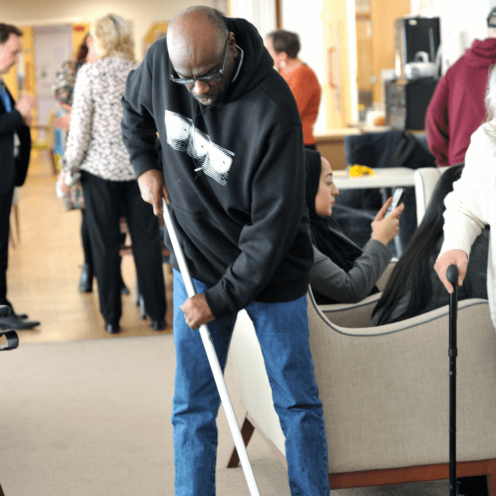 A man in a hoodie is sweeping the floor while others are sitting and standing in the background of a busy indoor space. - Home Instead
