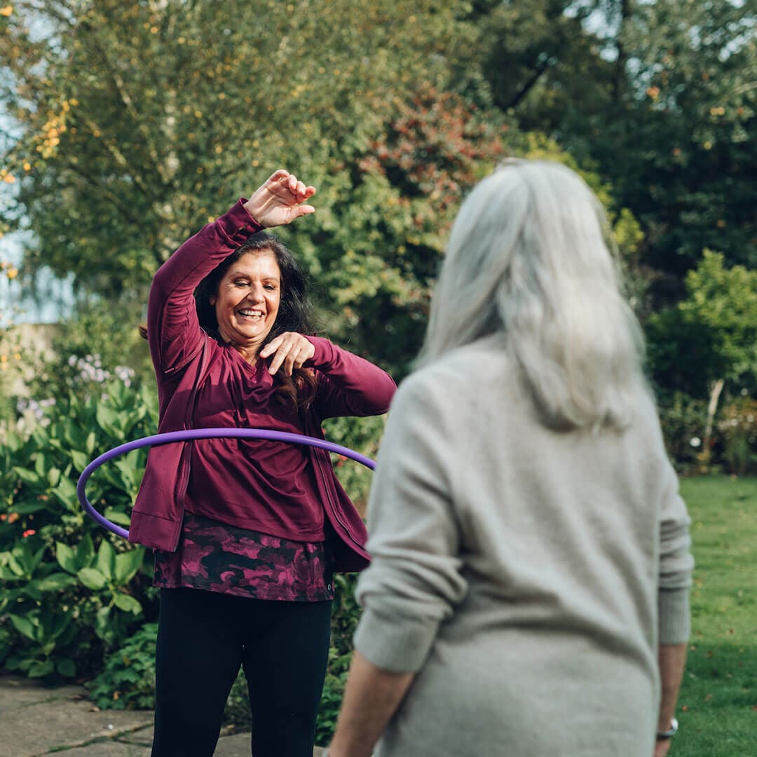 A caregiver hula-hoops outdoors while an older woman looks on, with greenery and trees in the background. - Home Instead