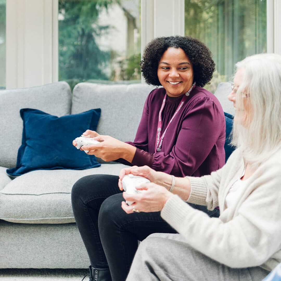 A younger woman and an older woman sit on a couch smiling and holding game controllers, engaging in a video game. - Home Instead