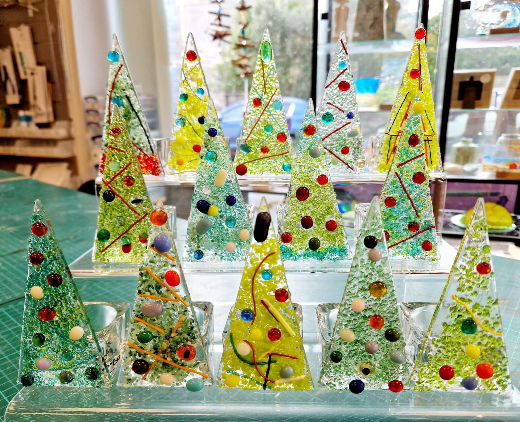A display of colorful, handcrafted glass Christmas trees adorned with beads and whimsical patterns on a table. - Home Instead