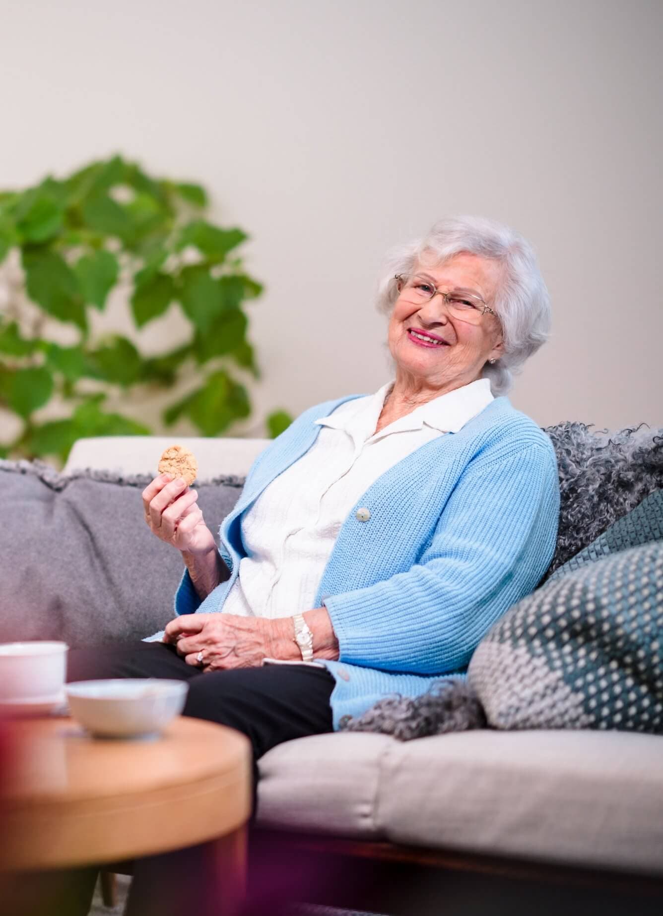 Elderly woman with white hair, wearing a blue cardigan, enjoys a cookie while sitting on a couch at home. - Home Instead