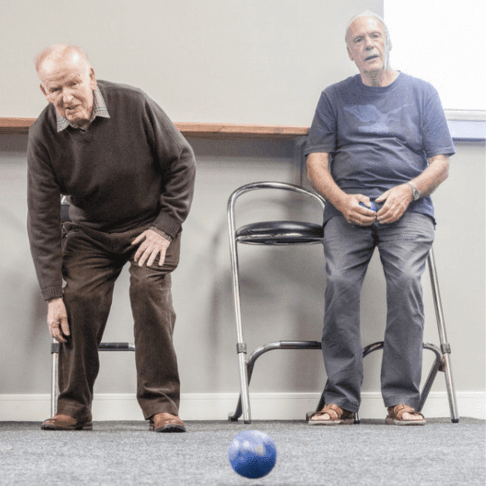Two elderly men play indoor bowls; one is bowling, while the other watches from a chair. - Home Instead
