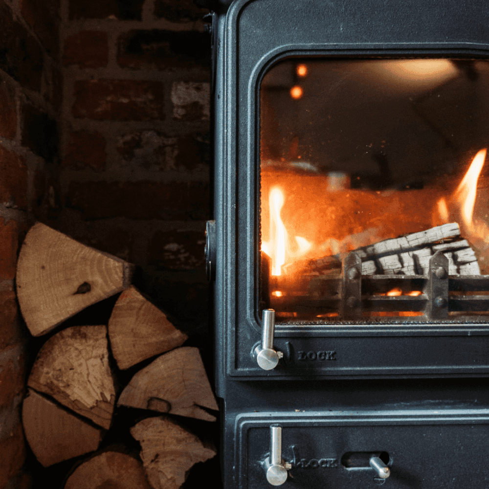 A black wood-burning stove with a fire inside, placed next to a stack of chopped wood on a brick wall. - Home Instead