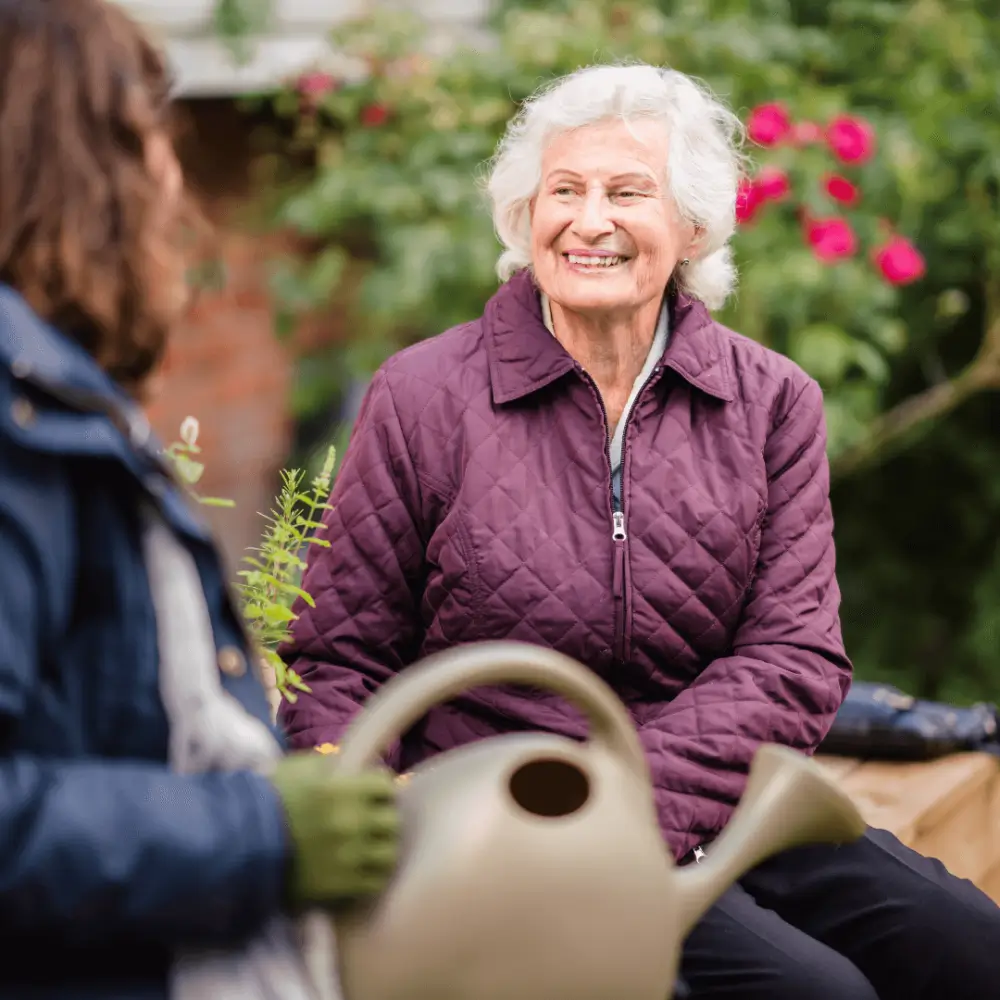 Elderly woman with white hair smiles, seated outside in a garden, conversing with another person holding a watering can. - Home Instead