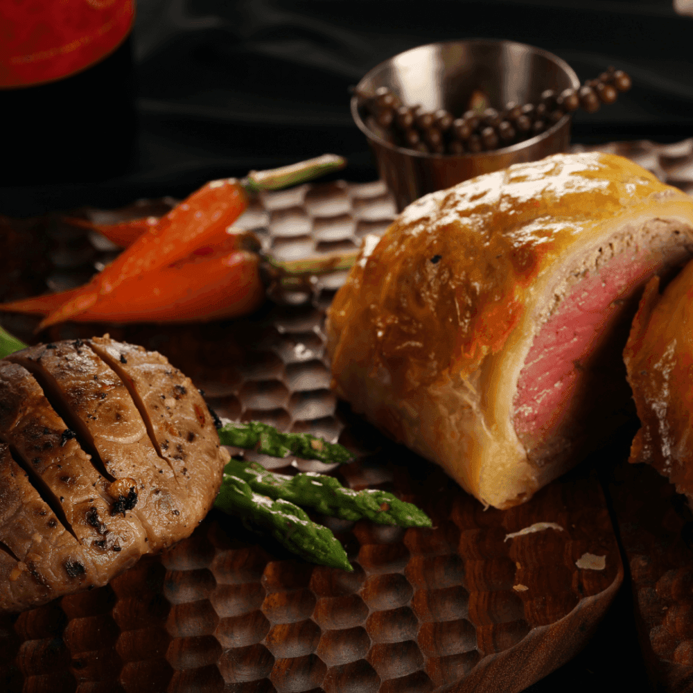 Beef Wellington, grilled mushroom, asparagus, and baby carrots on a wooden platter with a small bowl of peppercorns. - Home Instead