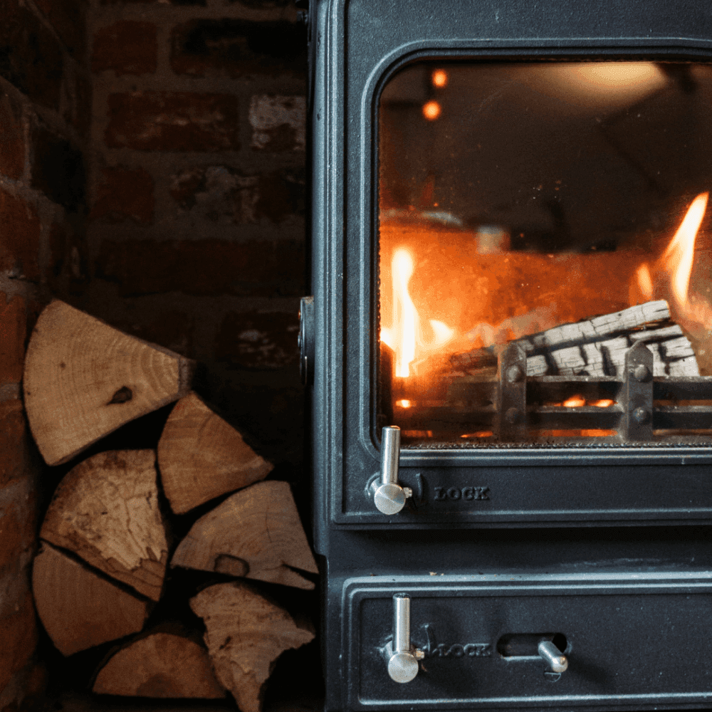 A cozy fireplace with logs burning, emitting warm flames and heat, stacked wood nearby against a brick wall. - Home Instead