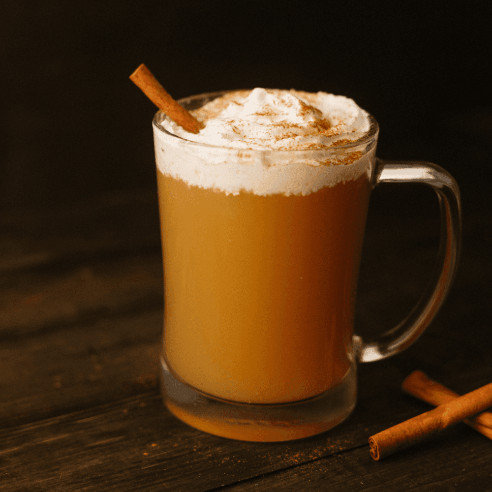 A mug of warm apple cider topped with whipped cream and a cinnamon stick on a dark wooden surface. - Home Instead