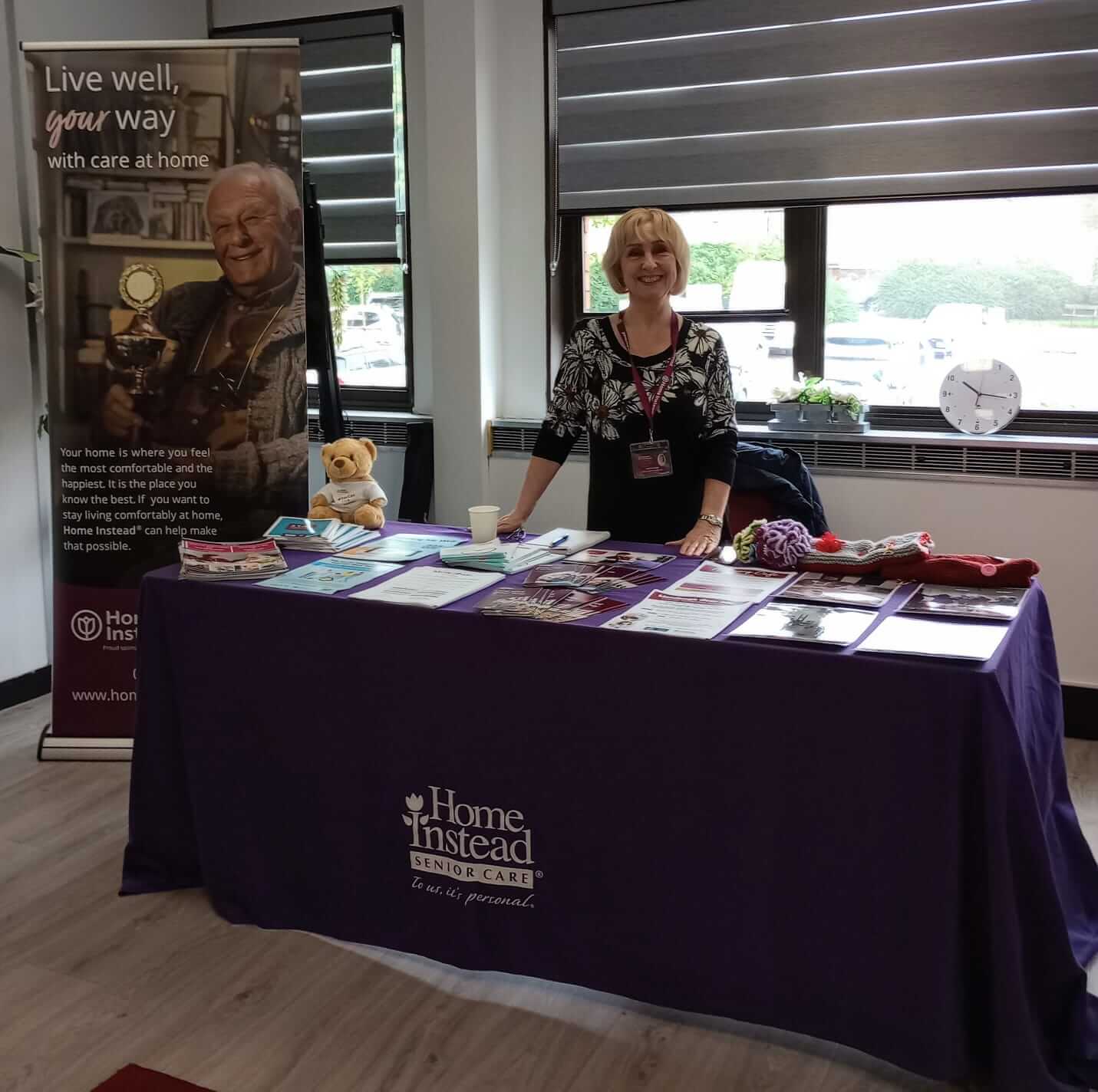 A woman stands behind a Home Instead Senior Care information table with brochures and a small teddy bear. - Home Instead