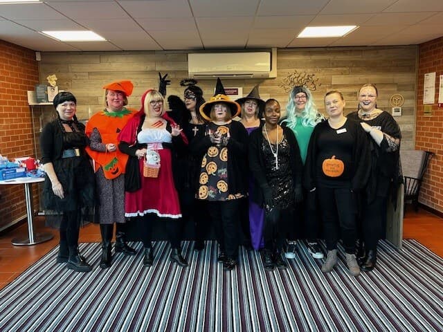 A group of people in various Halloween costumes stand together in a room with a striped rug. - Home Instead