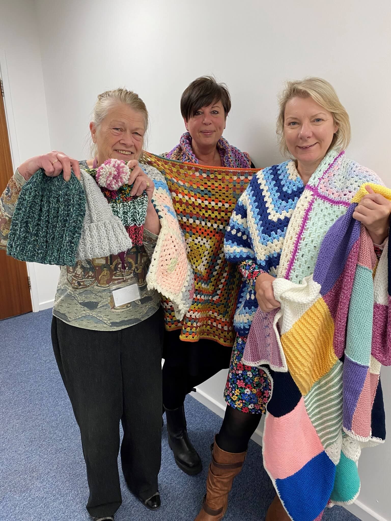 Three women holding and wearing colorful handmade crocheted items, including blankets and hats, in a room. - Home Instead