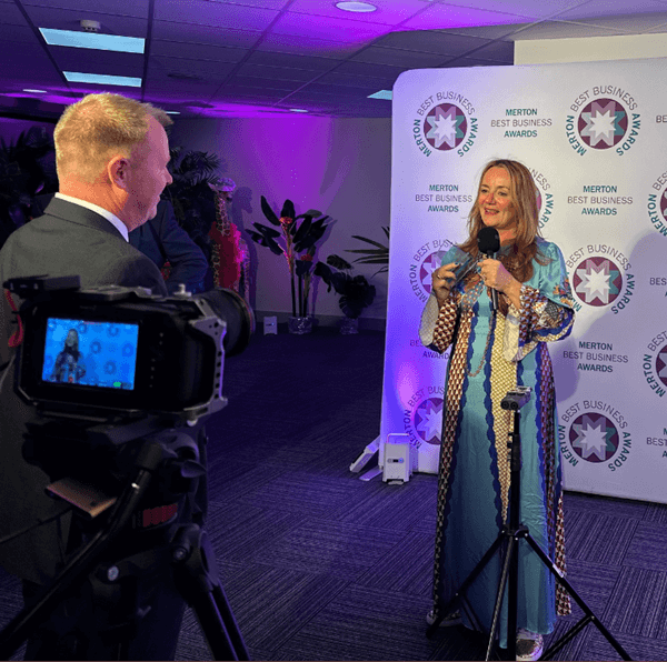 Woman in blue dress being interviewed on camera at Merton Best Business Awards backdrop. - Home Instead