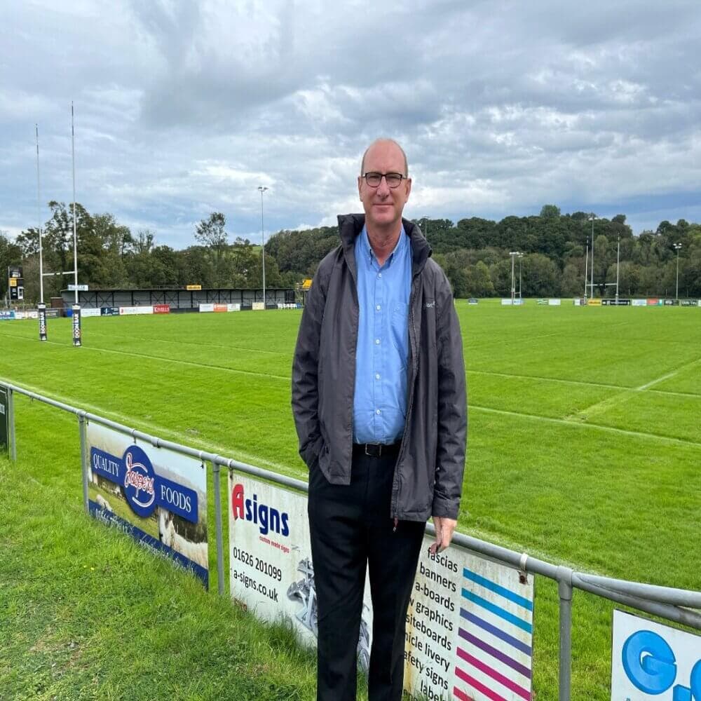 A man in glasses stands by a rugby field, wearing a gray jacket and blue shirt, with trees and cloudy sky in the background. - Home Instead