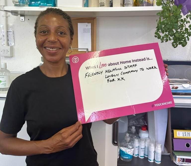 A person smiling, holding a sign that reads: "Friendly helpful staff. Lovely company to work for. xx". - Home Instead