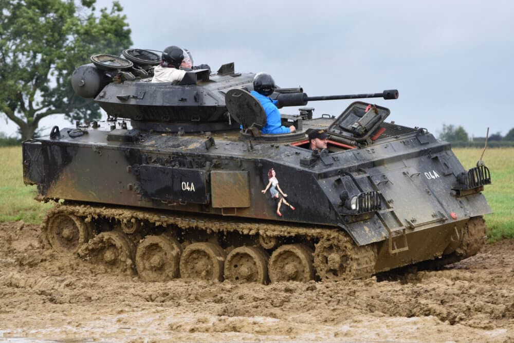 A military tank with people on top moving through a muddy field, with trees and cloudy sky in the background. - Home Instead