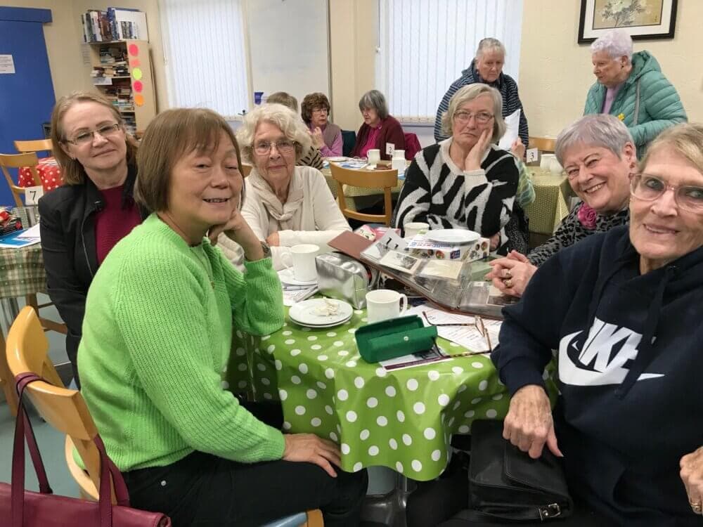 A group of elderly women sitting around a table with a green polka dot tablecloth, smiling and chatting in a cozy room. - Home Instead