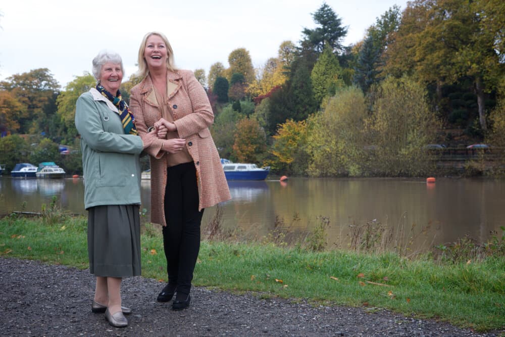 Two women laughing together by a serene riverside with boats and autumn trees in the background. - Home Instead