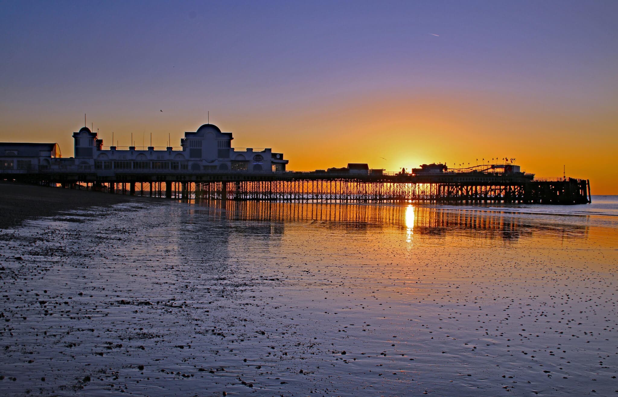 A pier extending into the ocean at sunset with its reflection visible on the calm water. - Home Instead