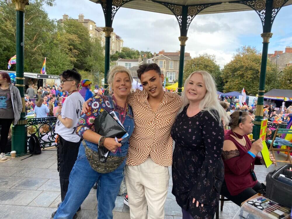 Three people posing together and smiling in front of a gazebo at an outdoor festival with a crowd in the background. - Home Instead